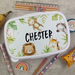 Puede incluir: Fiambrera blanca con dise&ntilde;o de animales de la selva. La fiambrera presenta un mono, una cebra, una jirafa, un le&oacute;n y un elefante, con el nombre "CHESTER" en negro. Hojas verdes y follaje selv&aacute;tico rodean a los animales. Tambi&eacute;n se ve una regla y &uacute;tiles escolares.