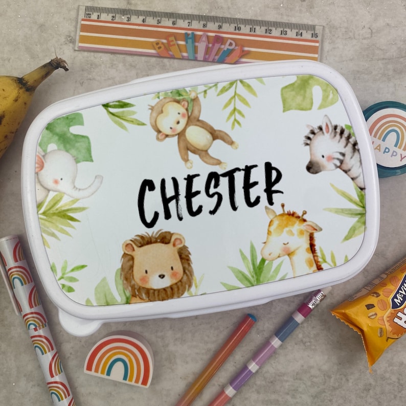 Puede incluir: Fiambrera blanca con dise&ntilde;o de animales de la selva. La fiambrera presenta un mono, una cebra, una jirafa, un le&oacute;n y un elefante, con el nombre "CHESTER" en negro. Hojas verdes y follaje selv&aacute;tico rodean a los animales. Tambi&eacute;n se ve una regla y &uacute;tiles escolares.