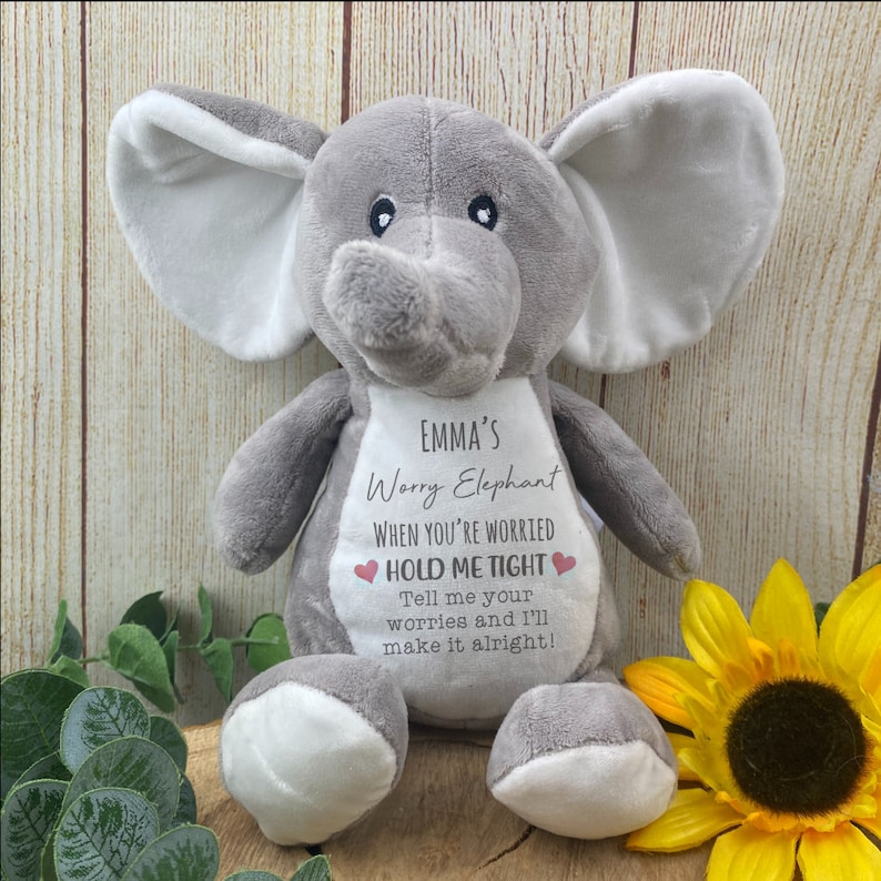 Op de afbeelding: Een grijze pluche olifant met witte accenten op de oren en voeten. De olifant heeft een slurf en kleine zwarte ogen. Tekst op de olifant luidt "EMMA'S Worry Elephant, HOLD ME TIGHT, Tell me your worries and I'll make it alright!"