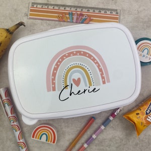 May include: White lunchbox with a rainbow design and the name "Cherie" in black script. The rainbow features pink, yellow, and blue arches with a heart at the center. Other school supplies include a ruler, pens, and a pencil sharpener.