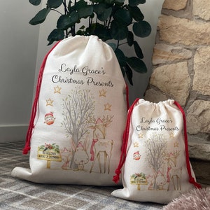 May include: Two beige Christmas gift bags with red drawstring closures. The bags feature a woodland scene with a deer, rabbit, and fox, along with the text "Layla Grace's Christmas Presents". A sign reads "Do Not Open Before 25 December".