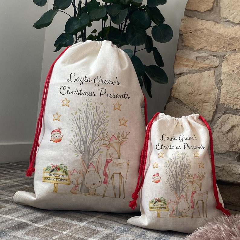 May include: Two beige Christmas gift bags with red drawstring closures. The bags feature a woodland scene with a deer, rabbit, and fox, along with the text "Layla Grace's Christmas Presents". A sign reads "Do Not Open Before 25 December".
