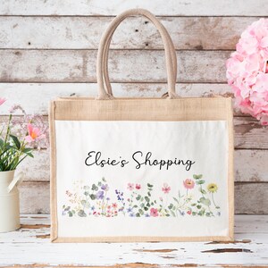 Puede incluir: Una bolsa de mano de yute natural con un inserto de lona blanca. La bolsa presenta un dise&ntilde;o floral con efecto acuarela y el texto "Elsie's Shopping".