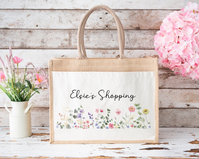 Puede incluir: Una bolsa de mano de yute natural con un inserto de lona blanca. La bolsa presenta un dise&ntilde;o floral con efecto acuarela y el texto "Elsie's Shopping".