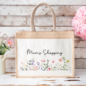 Puede incluir: Una bolsa de tela de yute natural con un inserto de lona blanca. La bolsa tiene un dise&ntilde;o floral y el texto "Mum's Shopping" impreso en la parte delantera.