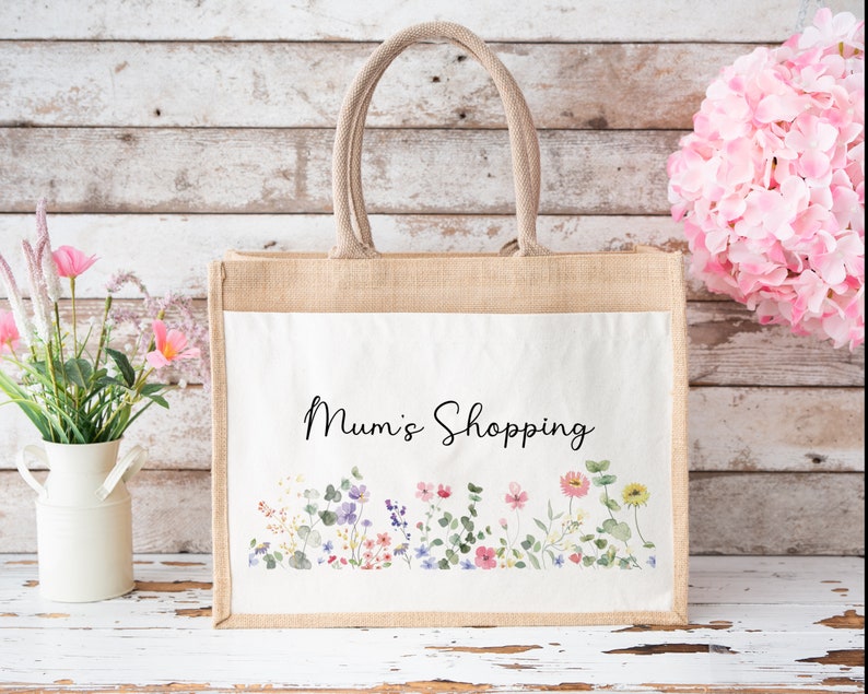 Puede incluir: Una bolsa de tela de yute natural con un inserto de lona blanca. La bolsa tiene un dise&ntilde;o floral y el texto "Mum's Shopping" impreso en la parte delantera.