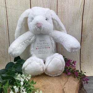 Op de afbeelding: Een witte pluche konijn met lange oren en een roze neus. Het konijn heeft een bedrukte tekst: "Tabitha's Worry Bunny" en "Hold Me Tight". Het staat op een houten oppervlak met bloemen.
