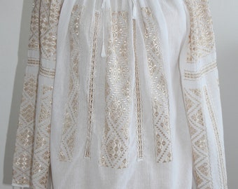 May include: White long-sleeved blouse with intricate gold embroidery. The blouse features a round neckline with a tie closure and tassels. The sleeves and front panels are adorned with detailed geometric and floral patterns.