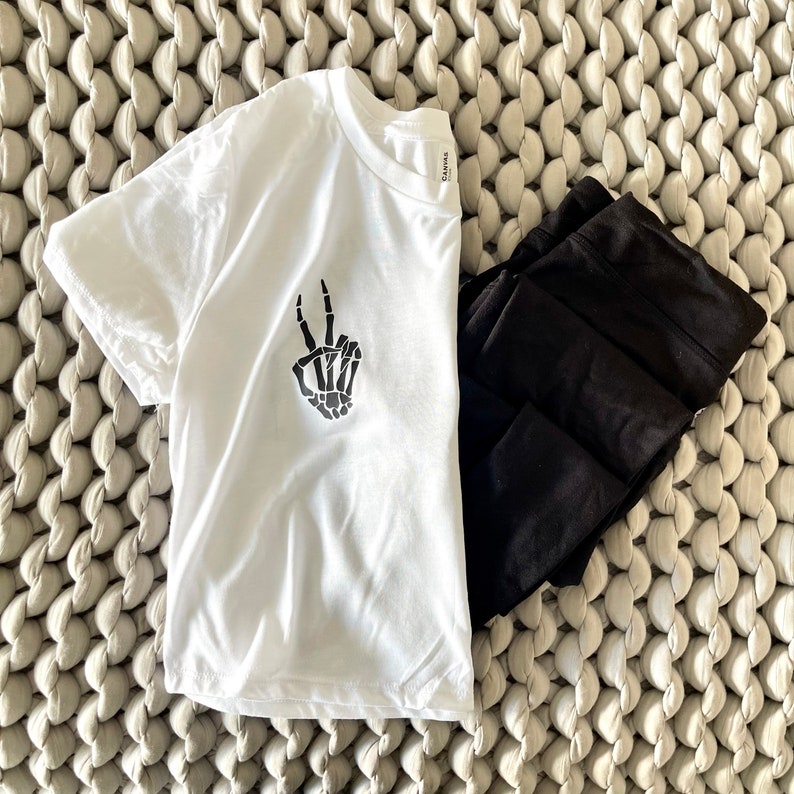 May include: White t-shirt with a black skeleton hand making a peace sign. The shirt is folded and lying on a white textured surface.