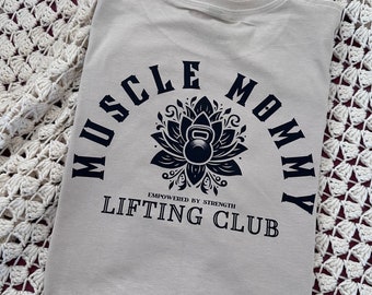 May include: A beige t-shirt with a black graphic design that says "Muscle Mommy Lifting Club" with a stylized lotus flower and a kettlebell in the center. The text "Empowered by Strength" is below the graphic.
