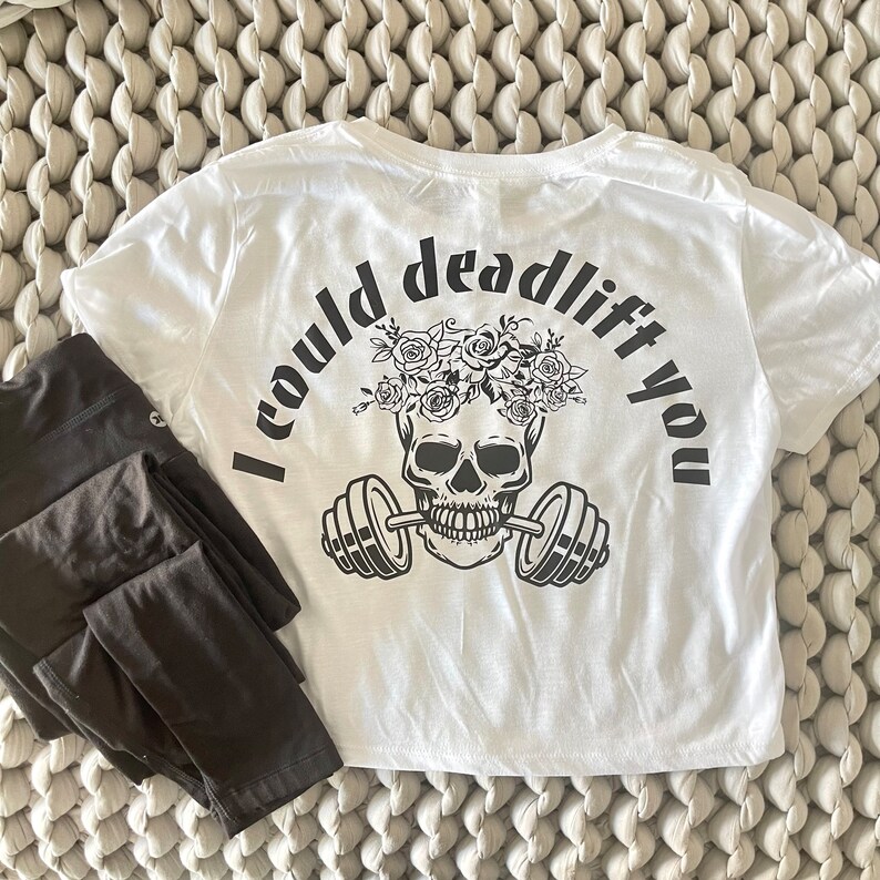 May include: White crop top with a black skull graphic wearing a flower crown and holding a barbell. The text "I could deadlift you" is printed on the back of the shirt.