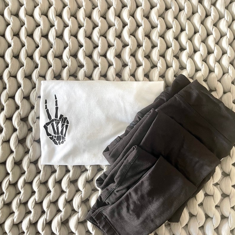May include: A white t-shirt with a black skeleton hand making a peace sign. The t-shirt is lying on a white textured surface with a stack of black leggings.