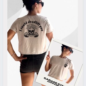 May include: A beige cropped t-shirt with a skull and barbell graphic on the back. The graphic says "I could deadlift you". The shirt is being worn by a person. The front of the shirt has a peace sign graphic. The shirt is from Garage Gym Apparel.