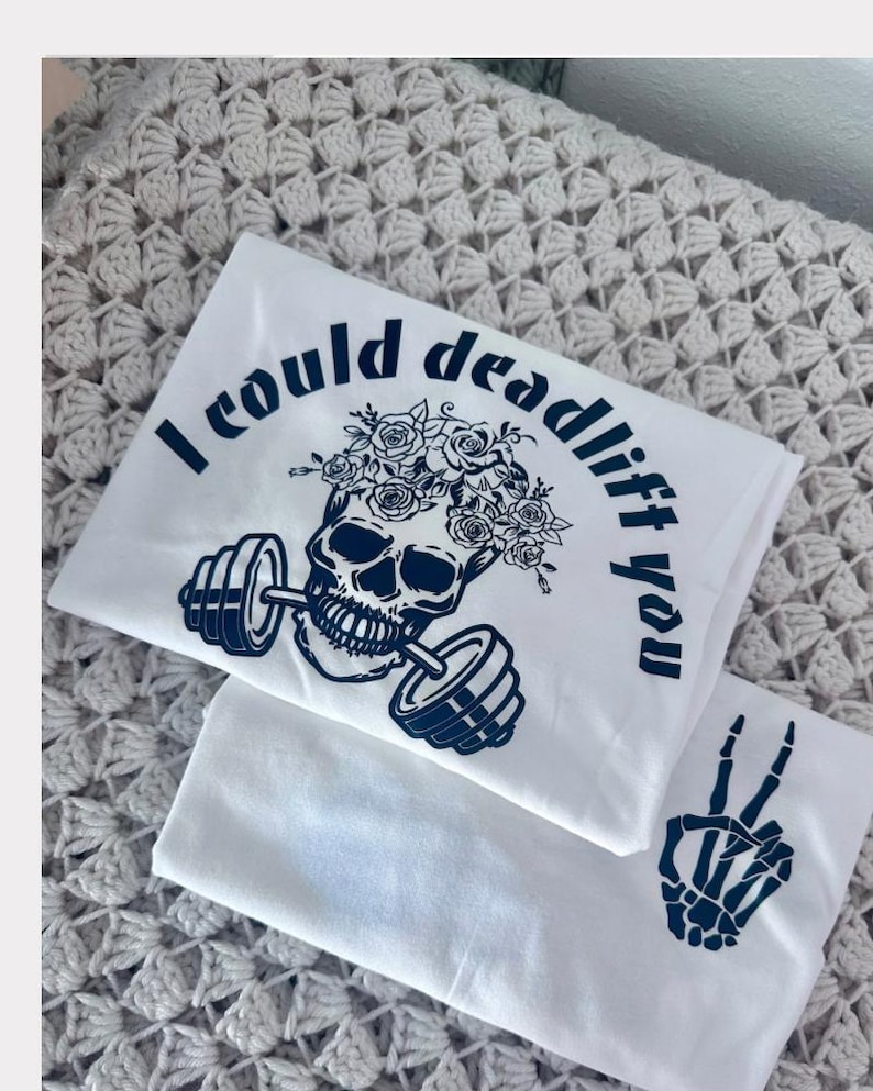 May include: White t-shirt with a blue skull graphic wearing a flower crown and holding a barbell. The text "I could deadlift you" is written around the skull. A second white t-shirt with a blue skeleton hand making a peace sign is visible in the background.