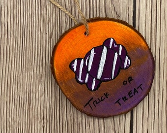 May include: A round wooden ornament with a purple and orange gradient background. A white and purple candy corn is painted on the ornament. The text "Trick or Treat" is written in black on the ornament.