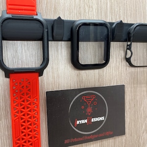 Könnte beinhalten: Drei 3D-gedruckte Uhrengehäuse, eines mit einem roten Band, eines mit einem klaren Displayschutz und eines mit einem durchgehenden schwarzen Gehäuse. Die Gehäuse sind an einem schwarzen Kunststoffhalter befestigt. Der Halter ist an einer Holzoberfläche befestigt. Eine Visitenkarte mit dem Text "RYAN DESIGNS 3D Printed Designs and Gifts" befindet sich in der unteren rechten Ecke des Bildes.