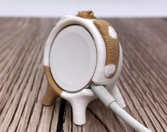 May include: White and brown 3D printed charging stand for an Apple Watch. The stand has a rounded shape and a white charging cable is plugged into the back.