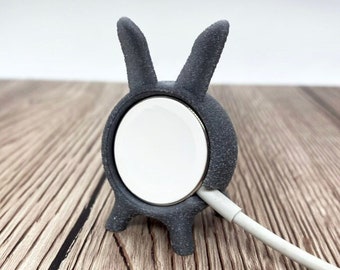 May include: A gray 3D printed rabbit-shaped stand for charging an Apple Watch. The stand has a cutout for the watch face and a hole for the charging cable.