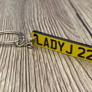 May include: A rectangular keyring with a yellow background and the text "LADY J 22" in black. It is attached to a silver key ring and chain. The background is a wooden surface.