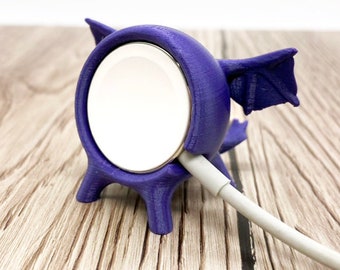 May include: Purple 3D printed dragon-shaped stand for an Apple Watch charging cable. The stand has a small hole for the charging cable to pass through and a larger hole to hold the watch.