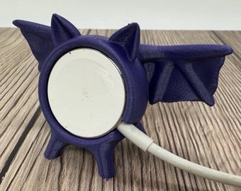 May include: A purple 3D printed bat-shaped phone stand with a white charging pad and a white charging cable.