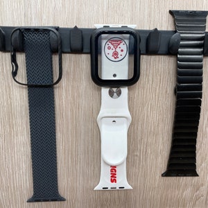 Könnte beinhalten: Drei Apple Watch-Armbänder hängen an einer schwarzen Wandhalterung. Die Armbänder sind ein schwarzes gewebtes Armband, ein weißes Sportarmband mit dem aufgedruckten Wort "SNPS" und ein schwarzes Milanese-Loop-Armband.