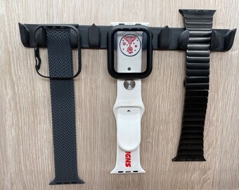 Könnte beinhalten: Drei Apple Watch-Armbänder hängen an einer schwarzen Wandhalterung. Die Armbänder sind ein schwarzes gewebtes Armband, ein weißes Sportarmband mit dem aufgedruckten Wort "SNPS" und ein schwarzes Milanese-Loop-Armband.