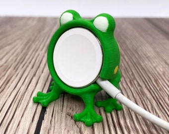 May include: A green frog-shaped charging stand for an Apple Watch. The stand has a white charging circle and a white charging cable.
