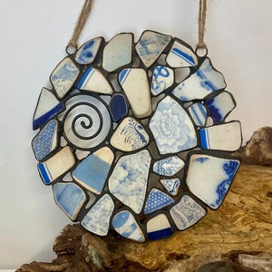 May include: A round wall hanging made of blue and white ceramic shards, arranged in a mosaic pattern. The design includes a central spiral and is suspended by a natural fiber cord. The ceramic pieces feature floral and geometric patterns.