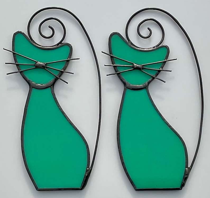 May include: Two teal blue stained glass cats with black wire outlines and curled tails. The cats have black wire whiskers and eyes.