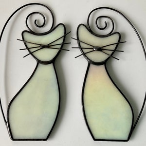 May include: Two white stained glass cats with black outlines and whiskers. The cats are facing forward and have a simple design.