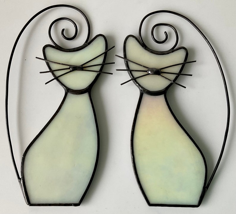 May include: Two white stained glass cats with black outlines and whiskers. The cats are facing forward and have a simple design.