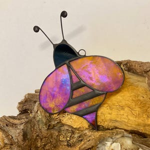 May include: A stained glass bee with iridescent wings in shades of pink, purple, and gold. The bee has a black body with horizontal stripes and delicate antennae. The artwork is mounted on a piece of driftwood.