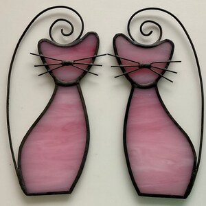 May include: Two pink stained glass cats with black outlines and wire whiskers. The cats are facing each other and have a curled tail.