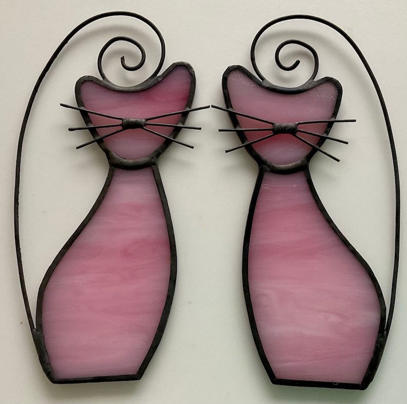 May include: Two pink stained glass cats with black outlines and wire whiskers. The cats are facing each other and have a curled tail.