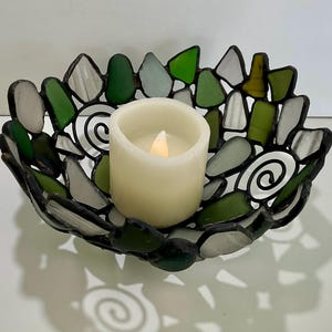 Puede incluir: Un portavelas decorativo con un diseño de vidrieras. El soporte presenta un mosaico de piezas de vidrio verdes, blancas y transparentes. Una vela blanca se encuentra en el centro. Los bordes del soporte son negros y proyecta una sombra en espiral.