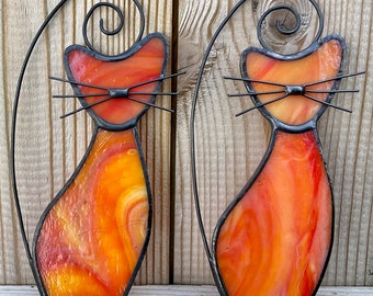 May include: Two stained glass cat silhouettes with black metal frames. The cats are orange and yellow with a marbled pattern. The cats are facing forward with their tails curled behind them.