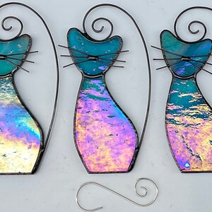 May include: Three iridescent glass cat ornaments with black wire frames. The cats are in a sitting position with their tails curled up behind them. The glass has a blue, purple, and pink iridescent finish.