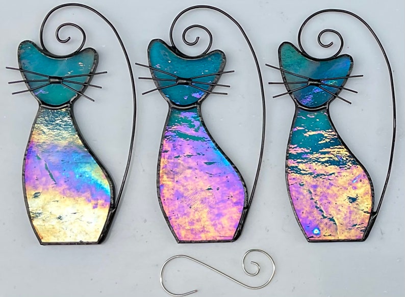May include: Three iridescent glass cat ornaments with black wire frames. The cats are in a sitting position with their tails curled up behind them. The glass has a blue, purple, and pink iridescent finish.