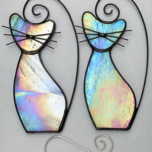 May include: Two iridescent glass cat ornaments with black wire frames and hanging loops. The cats are facing forward with their tails curled up behind them.