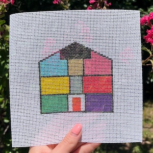 May include: A colorful cross-stitch pattern of a house with a patchwork design. The house features a black roof, blue, yellow, green, red, purple, and pink squares. The needlepoint is held up in front of a background of green foliage and pink flowers.