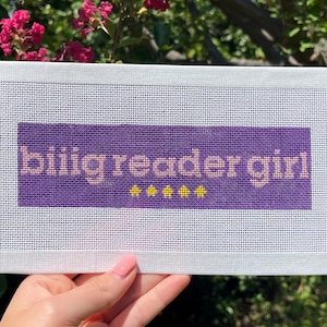 May include: A white needlepoint canvas with the text "biiig reader girl" in light purple and yellow. The canvas is held in a hand against a background of green leaves and pink flowers.