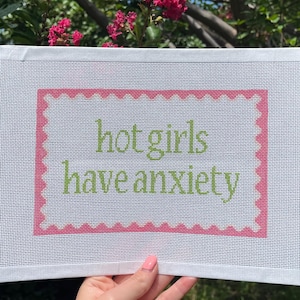 May include: A white canvas with a pink scalloped border and the text "hot girls have anxiety" in green. The canvas is held up in front of a background of green foliage and pink flowers.