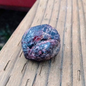May include: A small, irregularly shaped stone with a dark, textured surface. The stone has a mix of dark blue, burgundy, and reddish-pink hues. It rests on a weathered wooden surface, with visible wood grain.