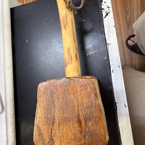 May include: A wooden mallet featuring a large, rectangular, stained brown head with visible wood grain and a cylindrical handle. A thin, weathered leather cord is tied around the upper portion of the handle, providing a simple loop for hanging or storage.