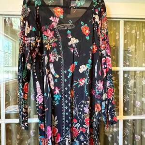 May include: A black long-sleeve tunic featuring a vibrant floral print with red, pink, orange, yellow, and teal flowers, leaves, and geometric patterns. The garment has a V-neckline, a relaxed fit, and a sheer, lightweight fabric construction.