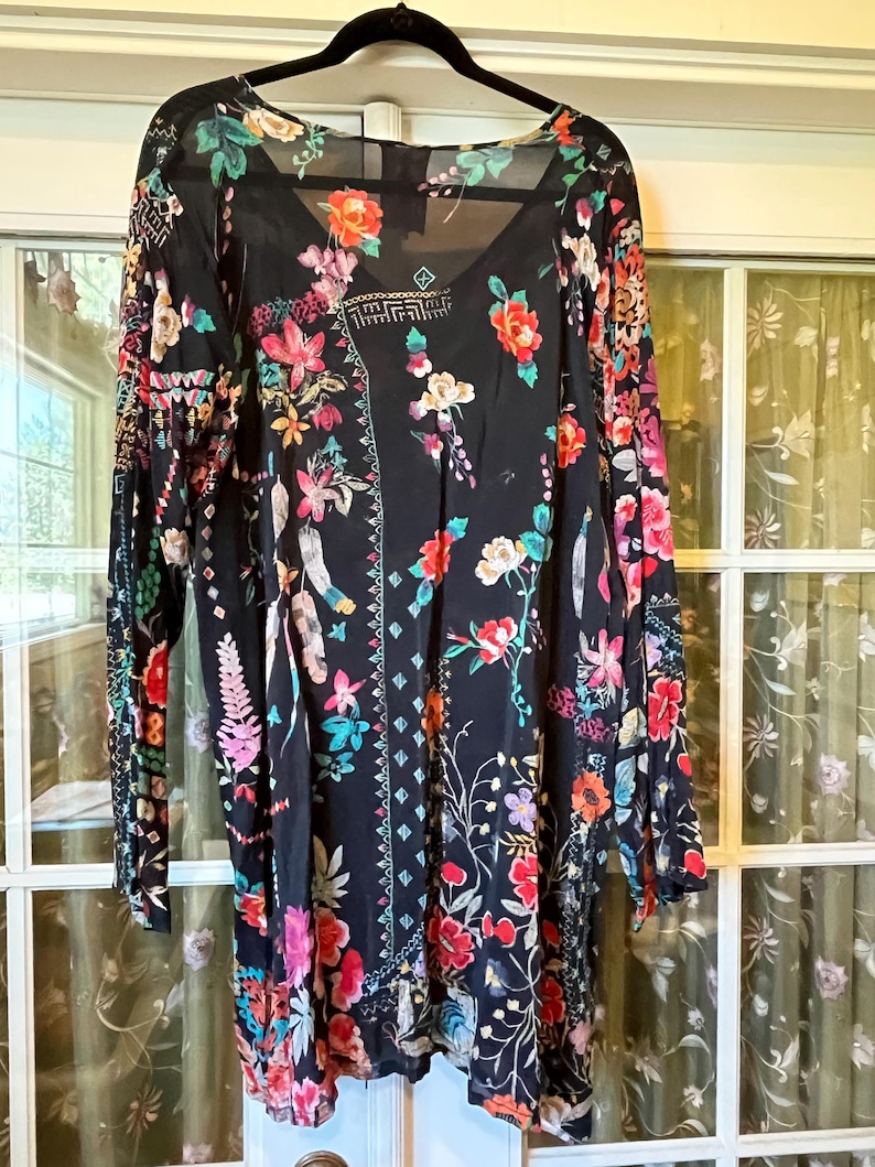 May include: A black long-sleeve tunic featuring a vibrant floral print with red, pink, orange, yellow, and teal flowers, leaves, and geometric patterns. The garment has a V-neckline, a relaxed fit, and a sheer, lightweight fabric construction.