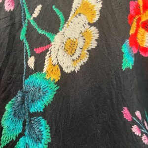 May include: A black fabric featuring vibrant floral embroidery. The design includes large white and yellow flowers, bright teal leaves, and smaller pink blossoms. The stitching is dense and textured, creating a colorful contrast against the dark background.