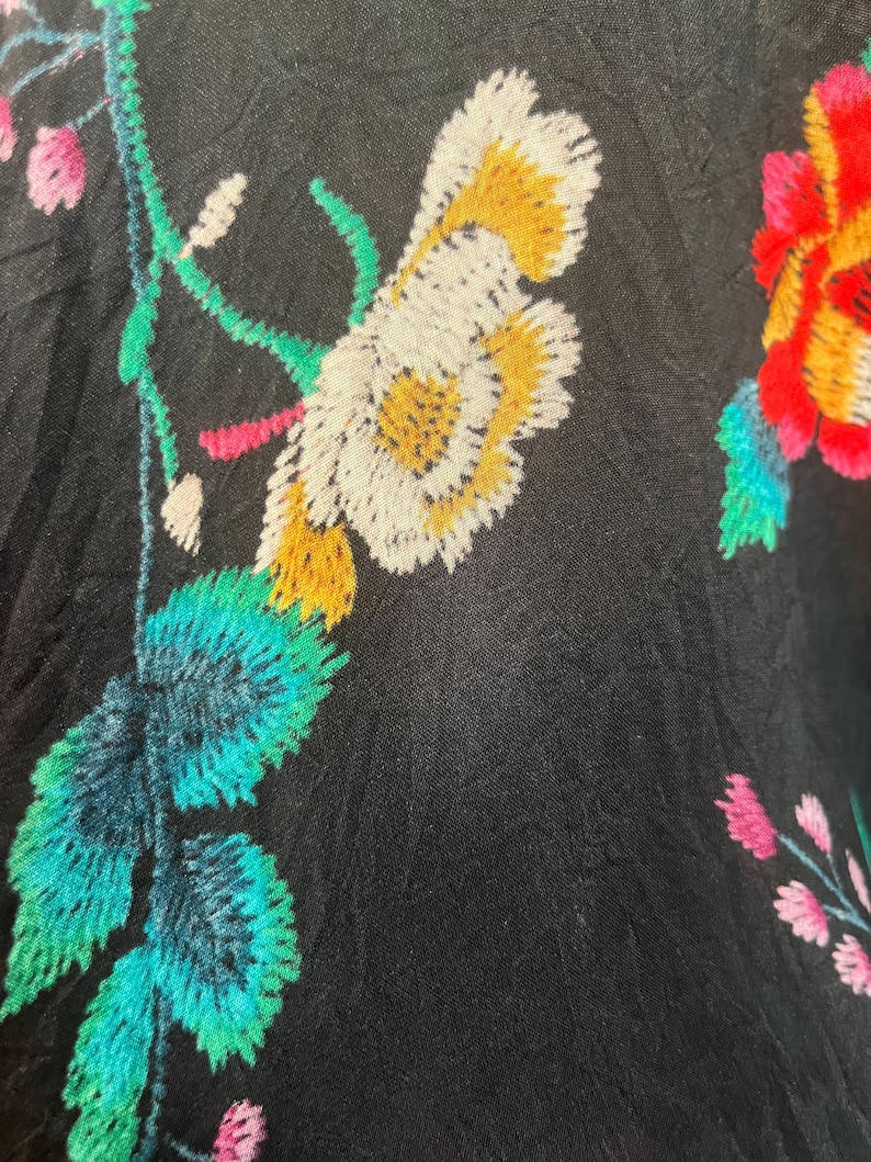 May include: A black fabric featuring vibrant floral embroidery. The design includes large white and yellow flowers, bright teal leaves, and smaller pink blossoms. The stitching is dense and textured, creating a colorful contrast against the dark background.