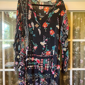 May include: A black long-sleeve tunic featuring a vibrant floral print with red, pink, and teal accents. The garment has a V-neckline and a label at the inner collar that reads Johnny Was. The fabric displays a mix of botanical and geometric patterns.